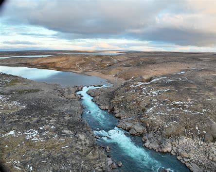 Aerial view of a blue river running through rocky tundra under a cloudy sky.